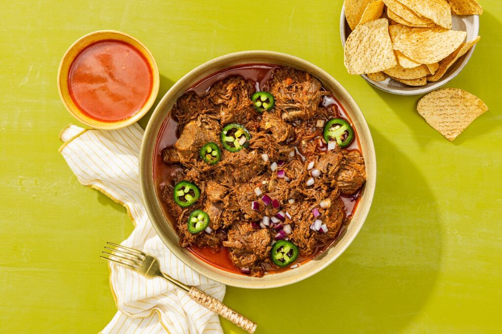 Beef birria in a bowl served with a side of birria sauce and tortilla chips on a green background. Explore food inspiration.
