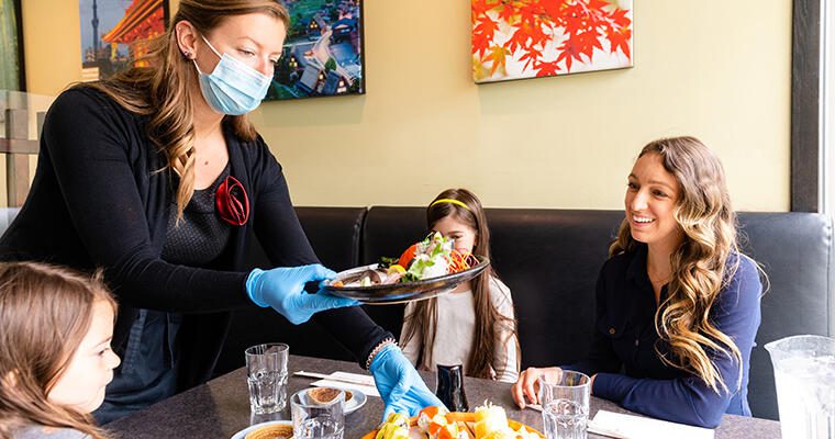 A server wearing a mask and gloves delivers plates of food to a restaurant table.
