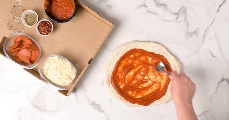 Hand spreading pizza sauce on dough with ingredients on side