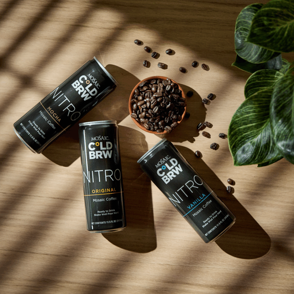 Cans of Mosaic Nitro Cold Brew laying down on a light wood surface with shadows being cast over them, laying near a bowl of coffee beans scattered over the table.