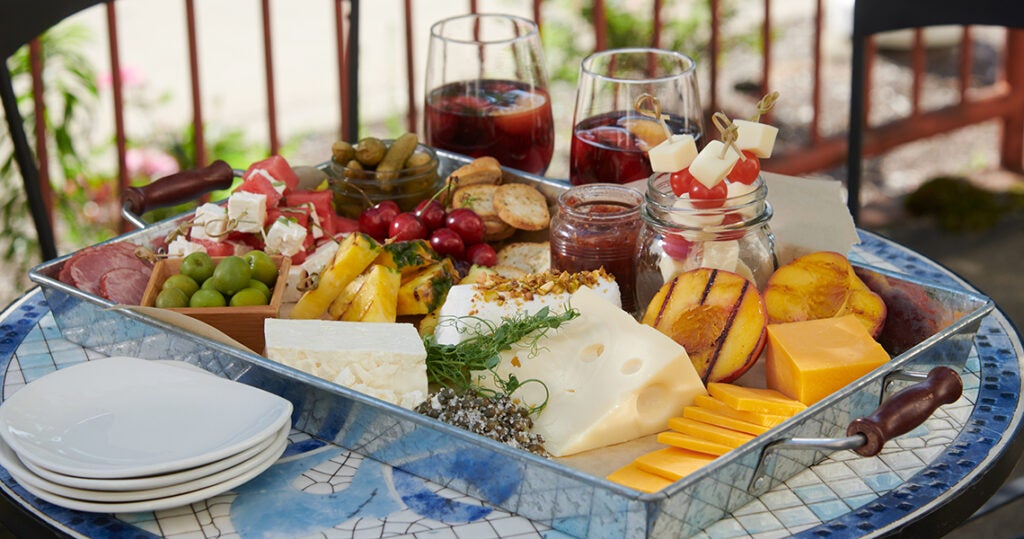 A Board that included cheese, fruits, crackers, pickles and has two drinks beside it.