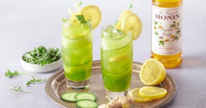 two glasses of lemonade and cucumber with a bottle of monin.