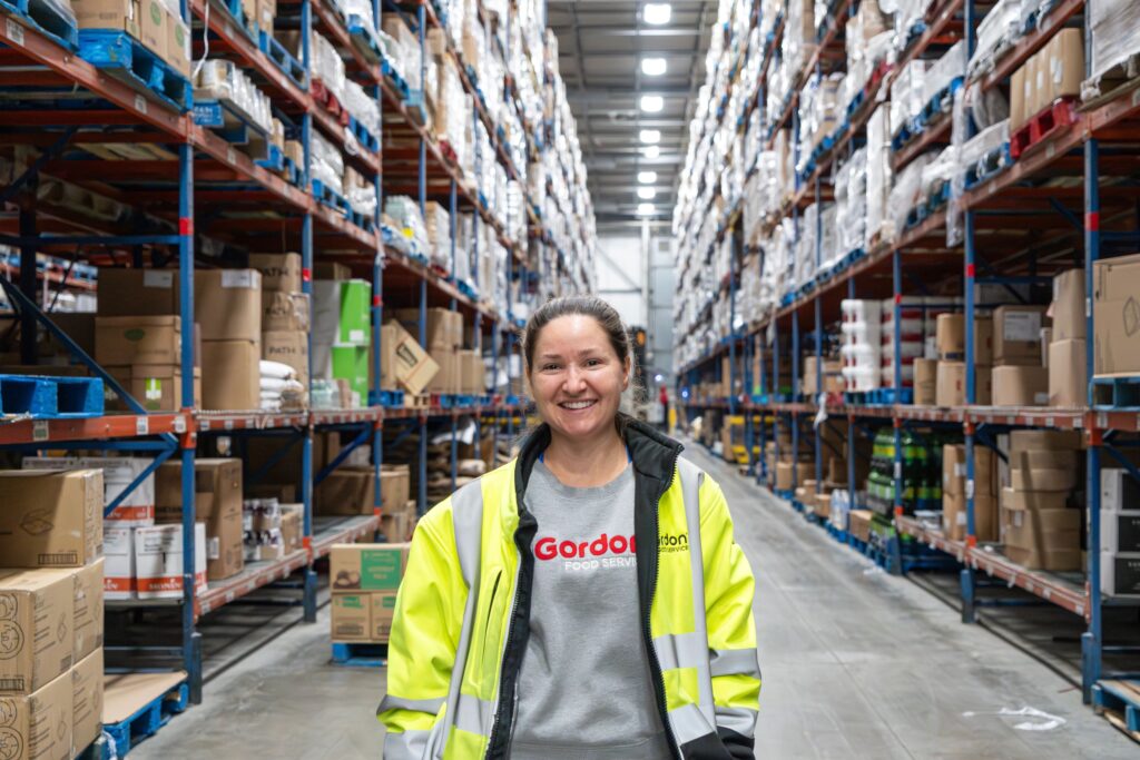 Warehouse employee smiling towards camera, wearing yellow safety jacket and company shirt