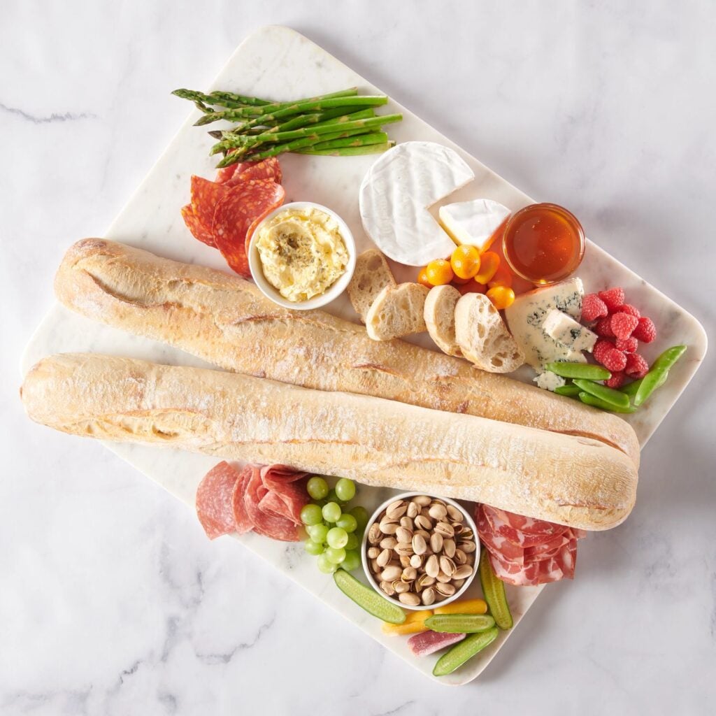 2 french baguettes surrounded by grazing board ingredients like meats, cheeses, and nuts.