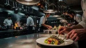 A busy commercial kitchen with four chefs preparing food, with the chef in the foreground plating a dish and other chefs working with various kitchen tools in the background.