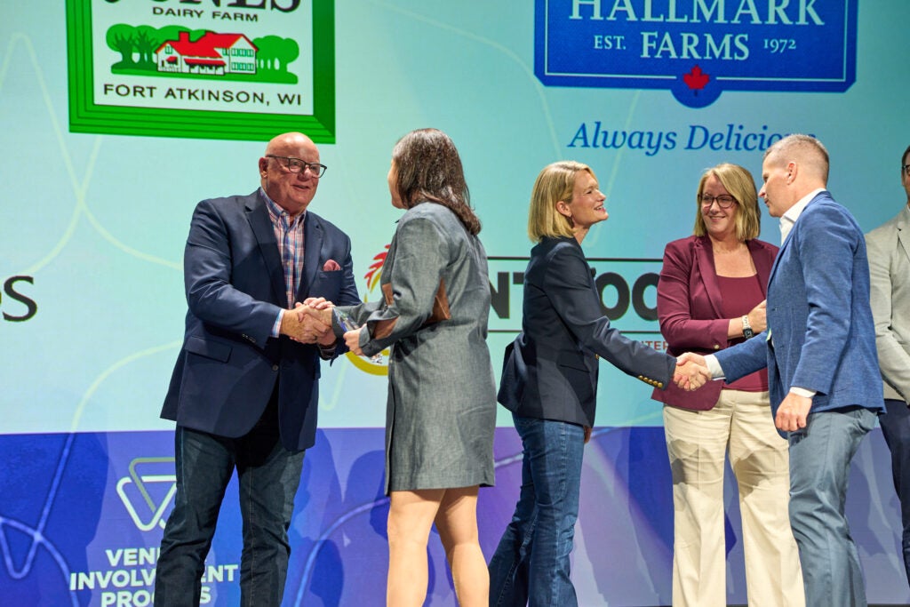 A diverse group of five people, including two men and three women, stand on a stage shaking hands, with a background featuring logos for Jones Dairy Farm and Hallmark Farms.