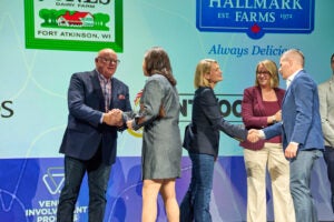 A diverse group of five people, including two men and three women, stand on a stage shaking hands, with a background featuring logos for Jones Dairy Farm and Hallmark Farms.