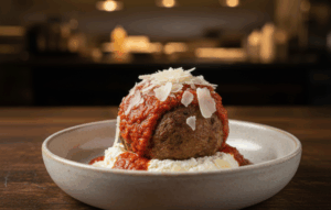 A savory meatball appetizer is presented on a white plate, featuring a large meatball topped with marinara sauce and shaved Parmesan flakes, resting on a bed of creamy ricotta cheese.