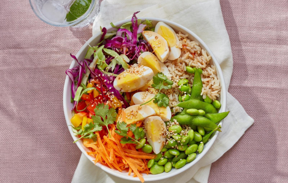 A vibrant, nutrient-dense bowl filled with hard-boiled eggs, edamame, shredded carrots, purple cabbage, and brown rice sits on a pink linen surface next to a glass of water with lime.