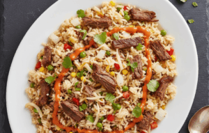 A vibrant platter of Birria Fried Rice featuring crispy beef chunks, mixed vegetables, and melted cheese, all drizzled with creamy sriracha and garnished with fresh cilantro.
