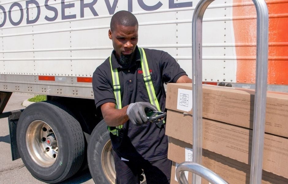 A delivery driver wearing a high-visibility vest and gloves uses a handheld electronic device to scan cardboard boxes stacked on a hand truck next to a foodservice delivery truck.