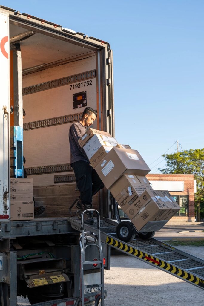 A delivery worker carefully guides a hand truck stacked high with cardboard boxes down a metal ramp from the back of an open delivery truck.