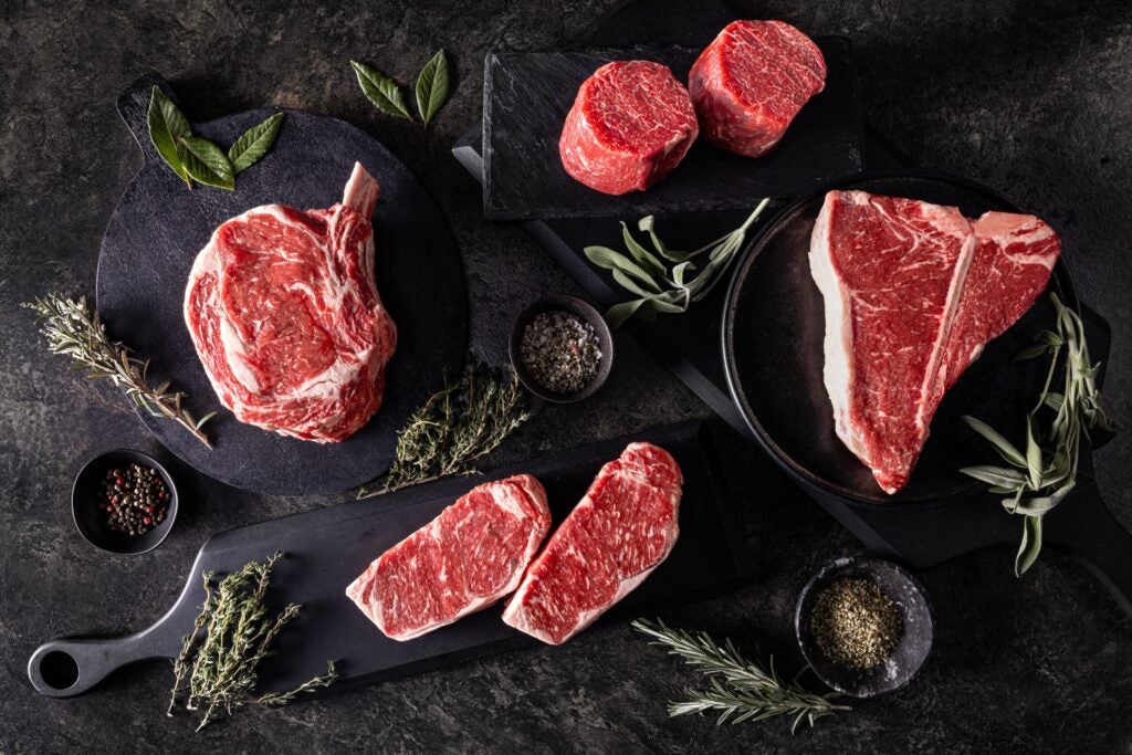 A selection of thick, raw marbled beef steaks, including ribeye, filet mignon, T-bone, and strip cuts, are artfully arranged on dark cutting boards and platters alongside fresh herbs and small bowls of spices.
