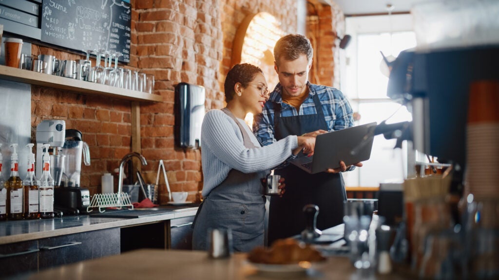 A woman and a man wearing aprons stand behind a coffee shop counter while looking together at a laptop held by the man.