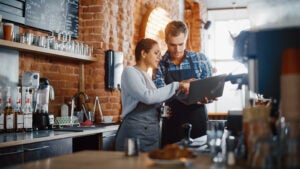 A woman and a man wearing aprons stand behind a coffee shop counter while looking together at a laptop held by the man.