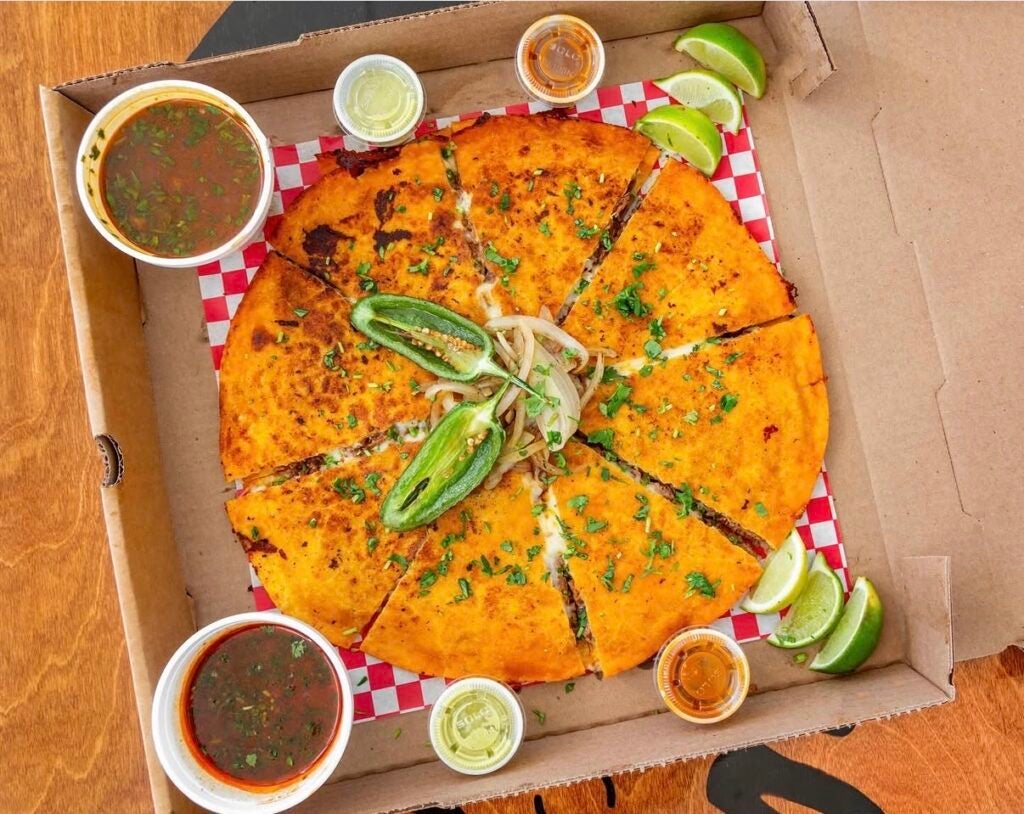 A sliced birria pizzadilla packed in a pizza box with consommé broth, salsas, jalapeños, and lime wedges.
