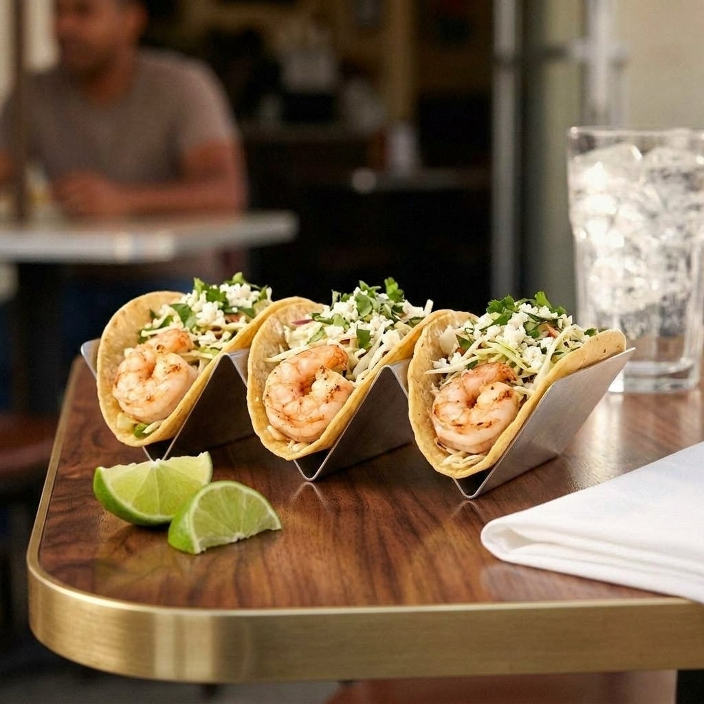 Three grilled shrimp tacos topped with slaw, cilantro, and crumbled cheese sit in a metal holder with lime wedges.
