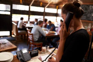 Una recepcionista de restaurante atiende una reserva por teléfono en el mostrador, con los comensales sentados al fondo.