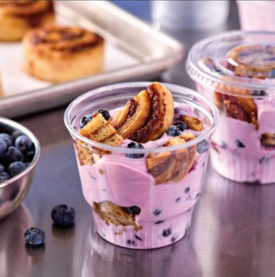 Blueberry cinnamon roll parfait layering purple yogurt, fresh blueberries, and cinnamon roll pieces in a clear plastic cup.