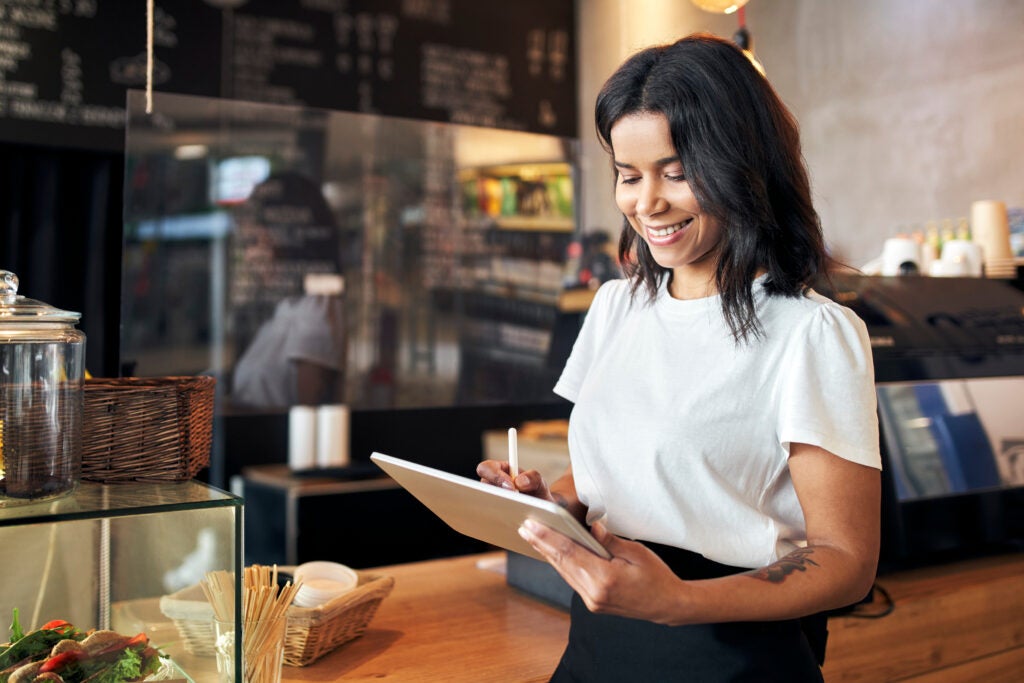 A smiling cafe worker writes on a tablet using a stylus behind the wooden counter.